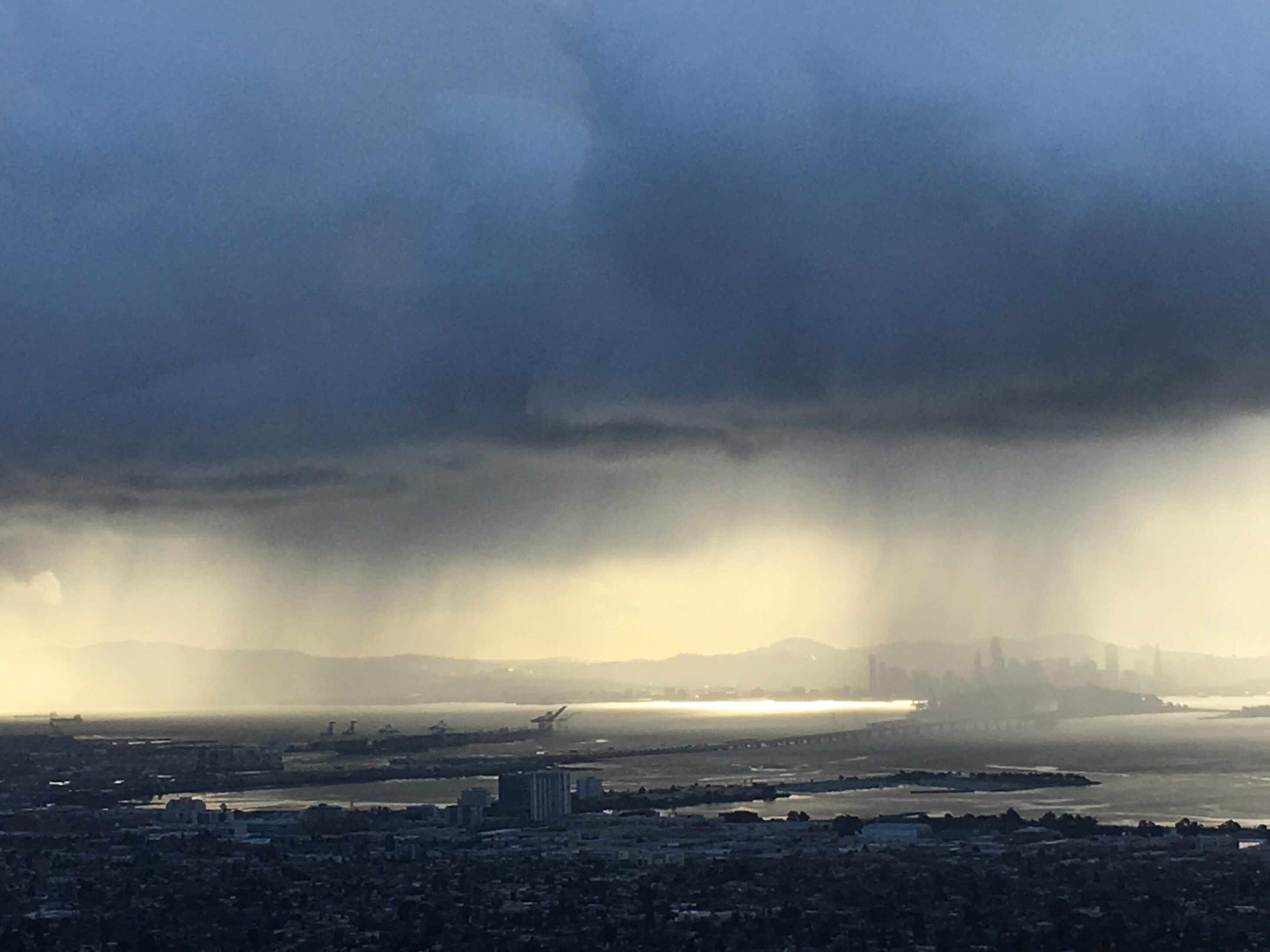 Storm clouds gather over a coastal city with rain falling on the water.